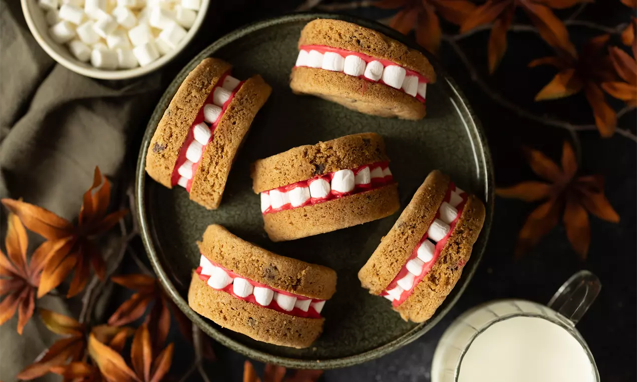 Biscuits dorés en forme de bouche farcis de crème rouge et mini-guimauves blanches, présentés sur une assiette verte avec des décorations automnales