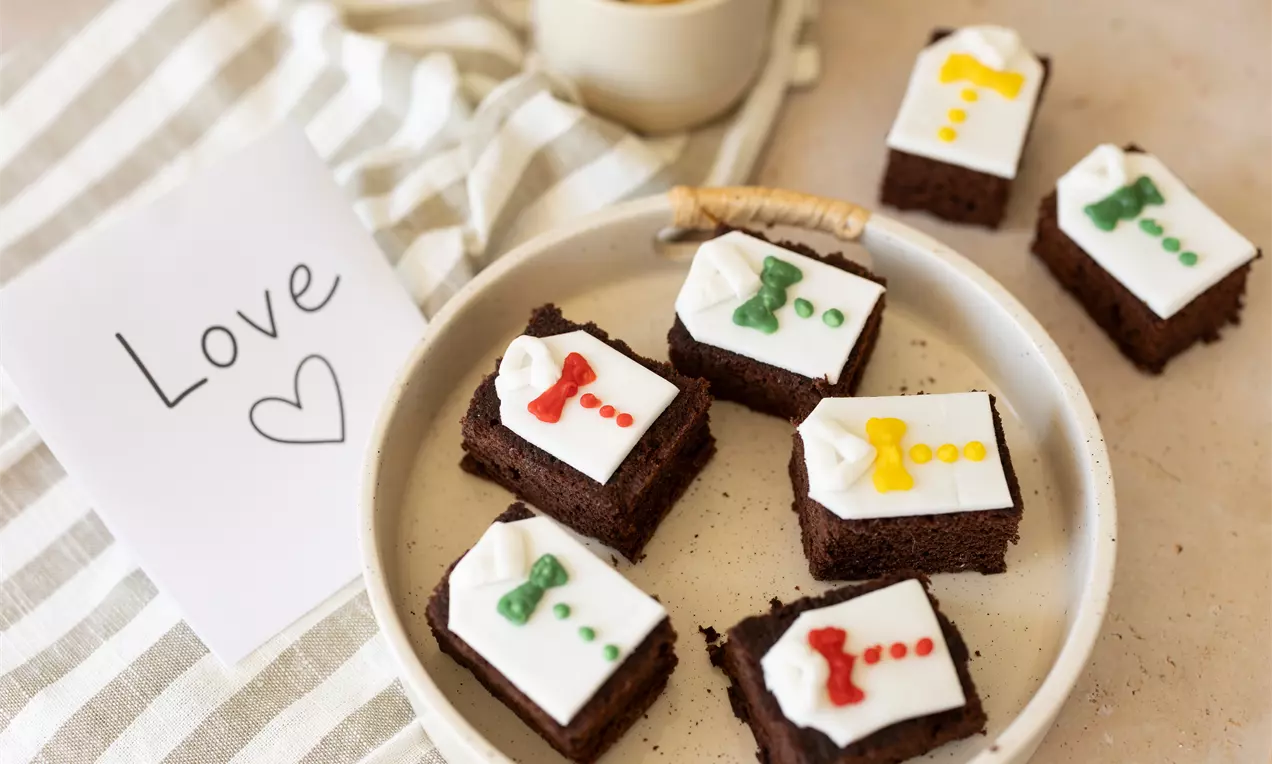 Chocoladevierkantjes met witte glazuurlaag, gedecoreerd met kleurrijke lijnen en strikken, geserveerd op een licht bord met een kaartje en koffie