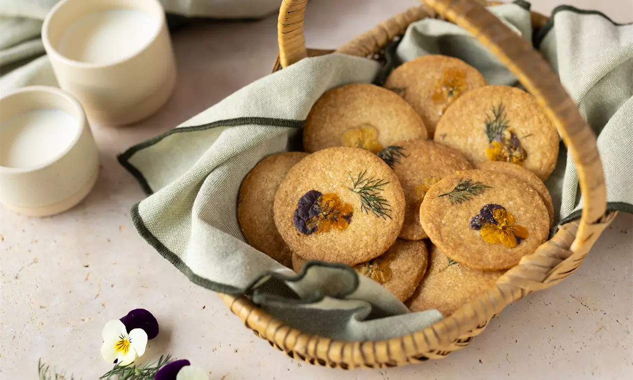 Goudbruine ronde koekjes met eetbare bloemen en dille, op een beige stoffen doek in een gevlochten mand, met melkglazen op de achtergrond