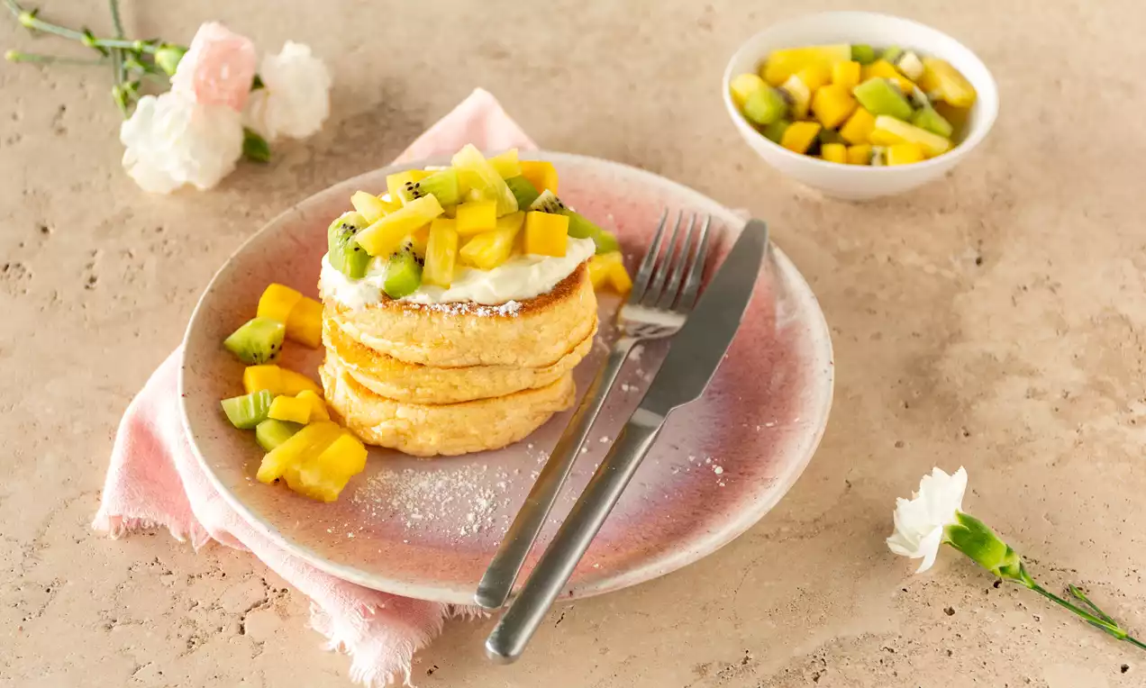 Gouden, luchtige pannenkoeken op een roze bord, bedekt met frisse stukjes mango en kiwi, een toefje romige witte yoghurt en een roze servet