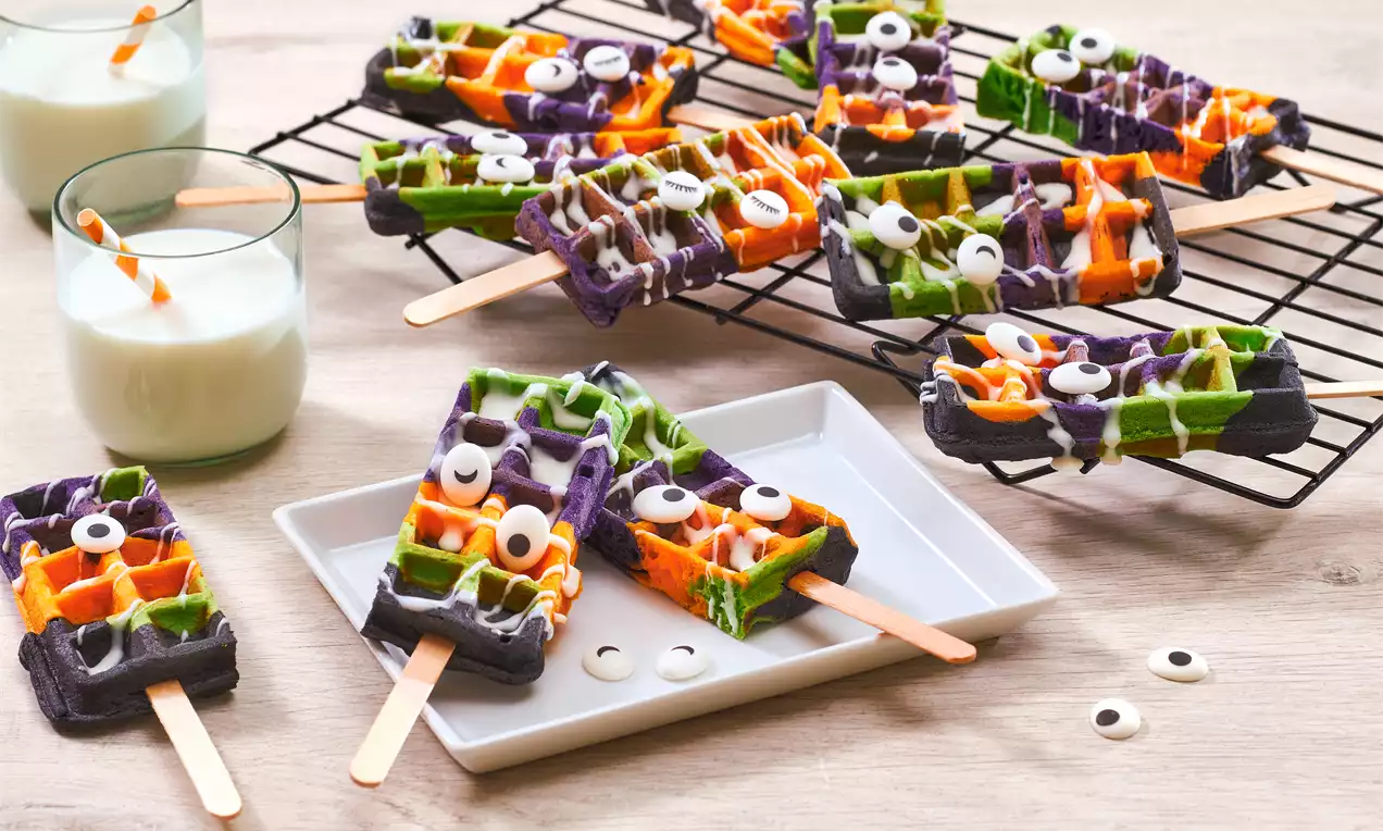 Halloween-wafels met oranje, groene en paarse kleuren, op houten stokjes, gegarneerd met witte glazuur en oogdecoraties op een rooster