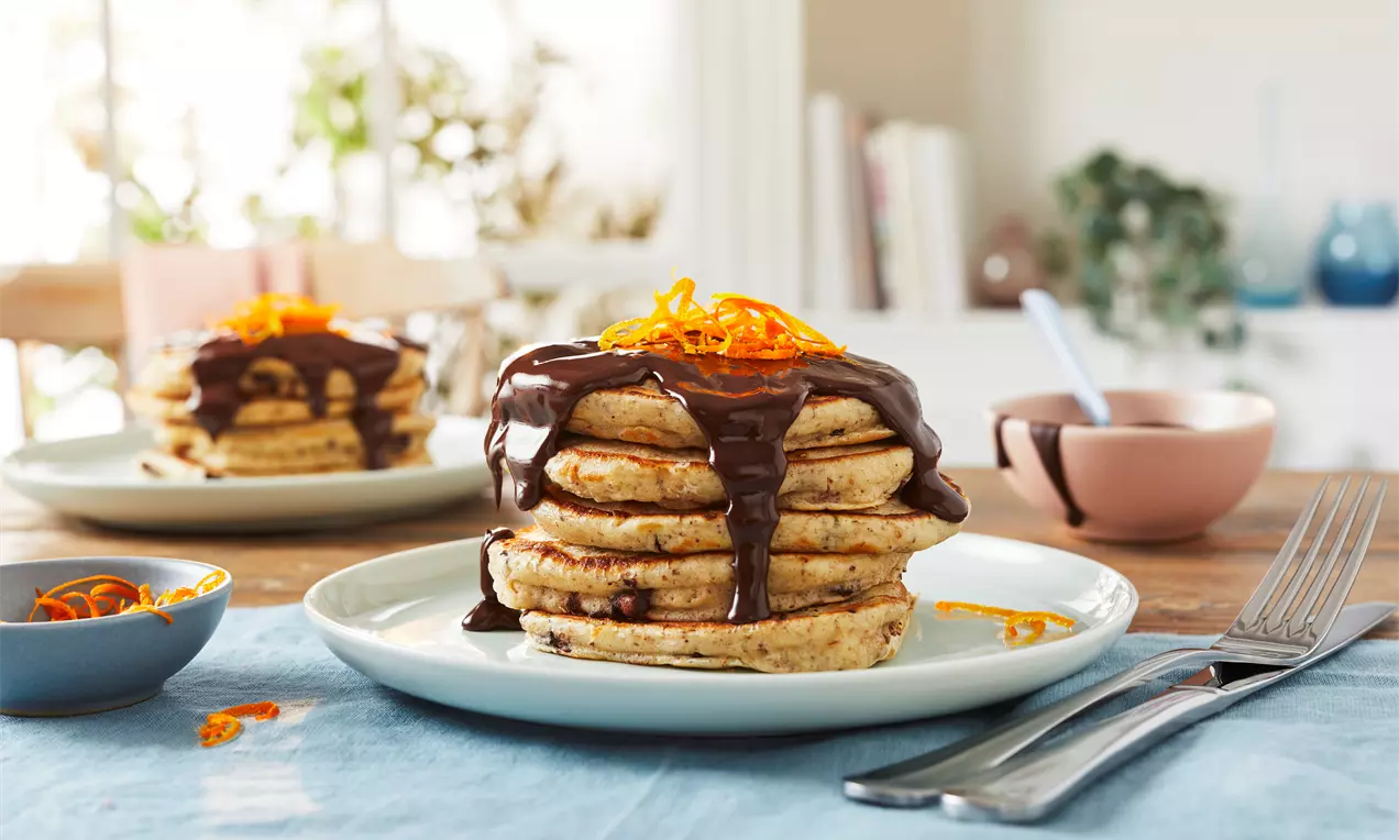 Een stapel goudbruine vegan pannenkoeken met glanzende chocoladesaus en feloranje sinaasappelschil op een lichtblauw bord