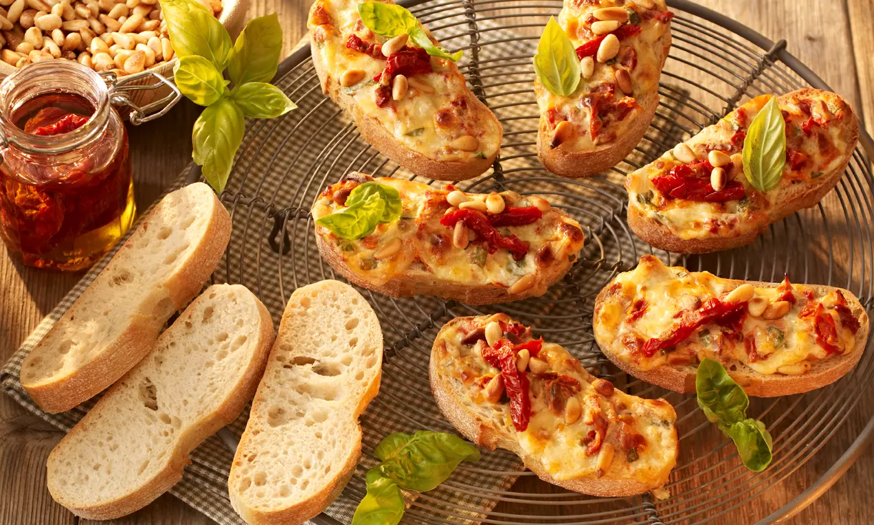 Tranches de ciabatta dorées, gratinées au fromage, garnies de basilic frais, tomates séchées et pignons, disposées sur une grille