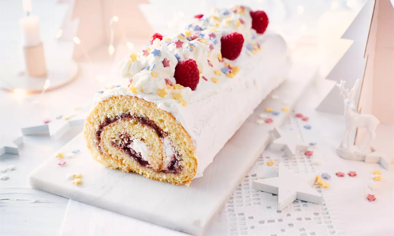 Bûche roulée à la génoise jaune pâle garnie de confiture rouge et crème blanche, décorée de framboises et étoiles colorées sur un plateau lumineux