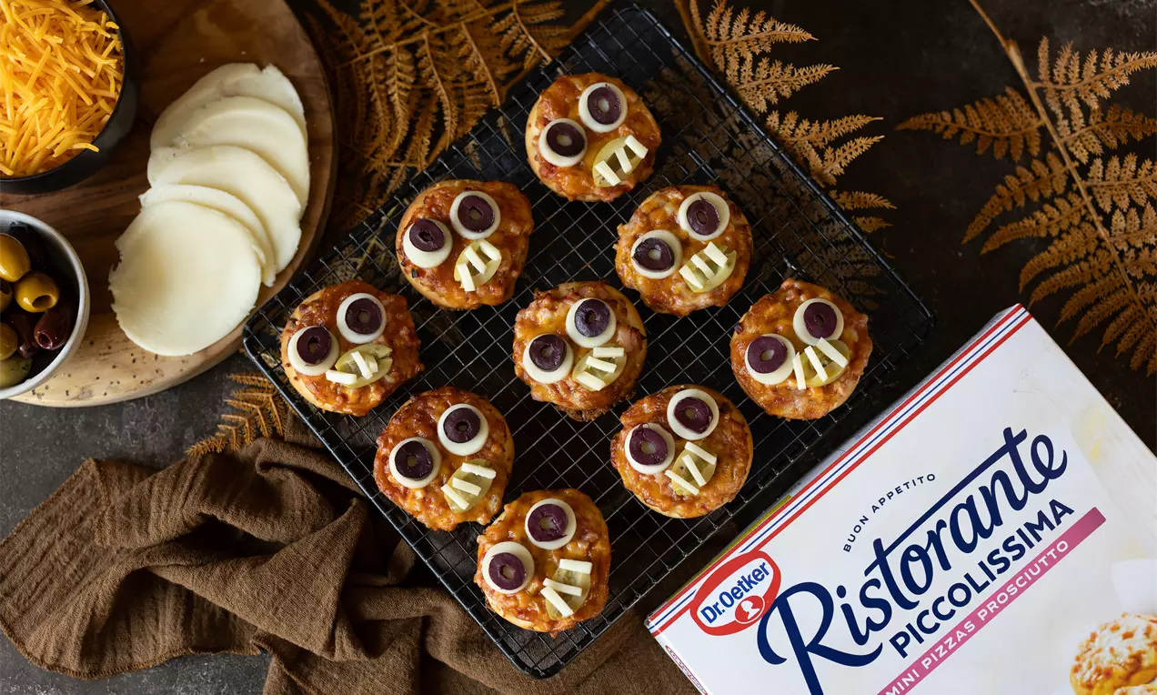 Mini-pizzas dorées en forme de têtes de squelettes avec olives noires, fromage blanc et jaune disposées sur une grille noire.