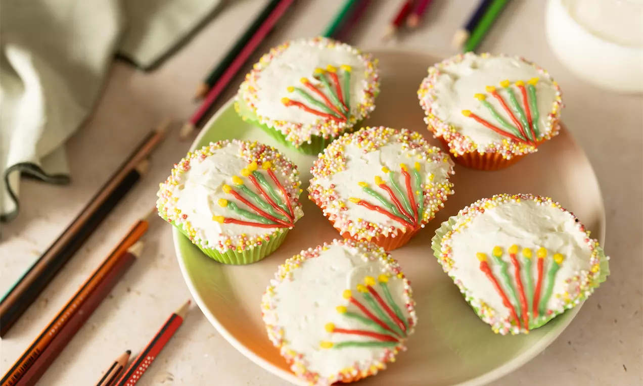 Zes cupcakes met witte glazuurlaag, kleurrijke sprinkels langs de randen en decoratieve lijnen in rood, geel en groen op een licht bord
