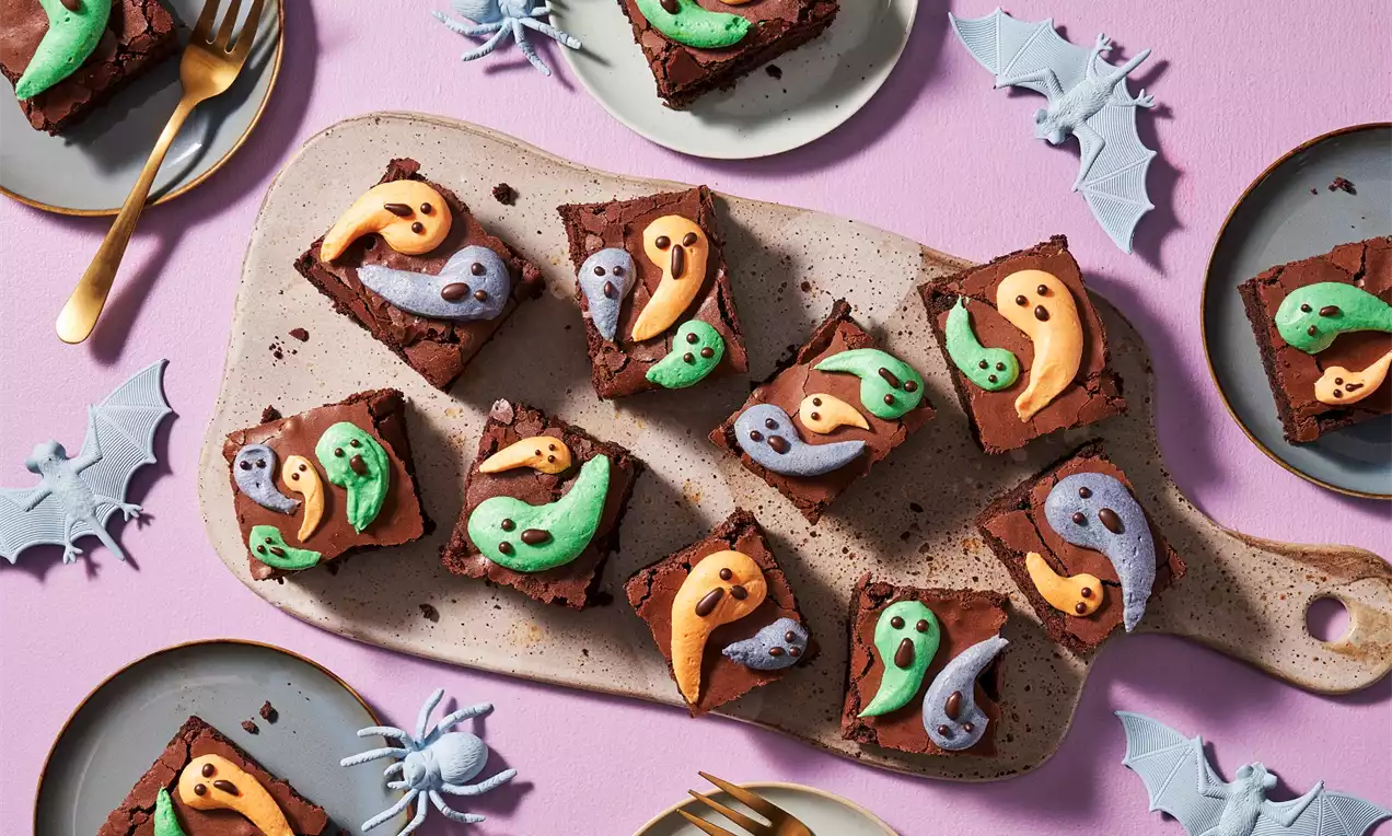 Brownies carrés moelleux au chocolat brun, décorés de fantômes colorés en glaçage orange, vert et violet, sur un plateau beige