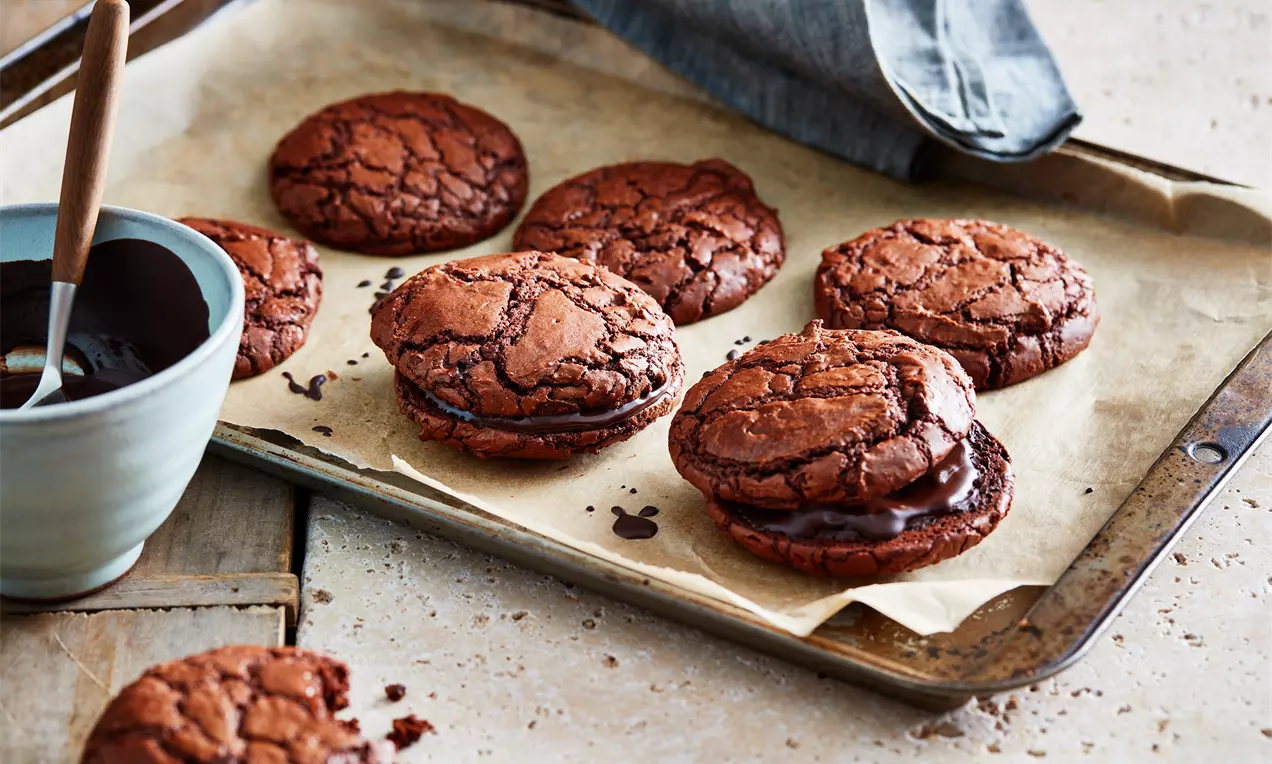 Cookies au chocolat brun foncé, aux bords légèrement craquelés, garnis de ganache, disposés sur une plaque recouverte de papier cuisson