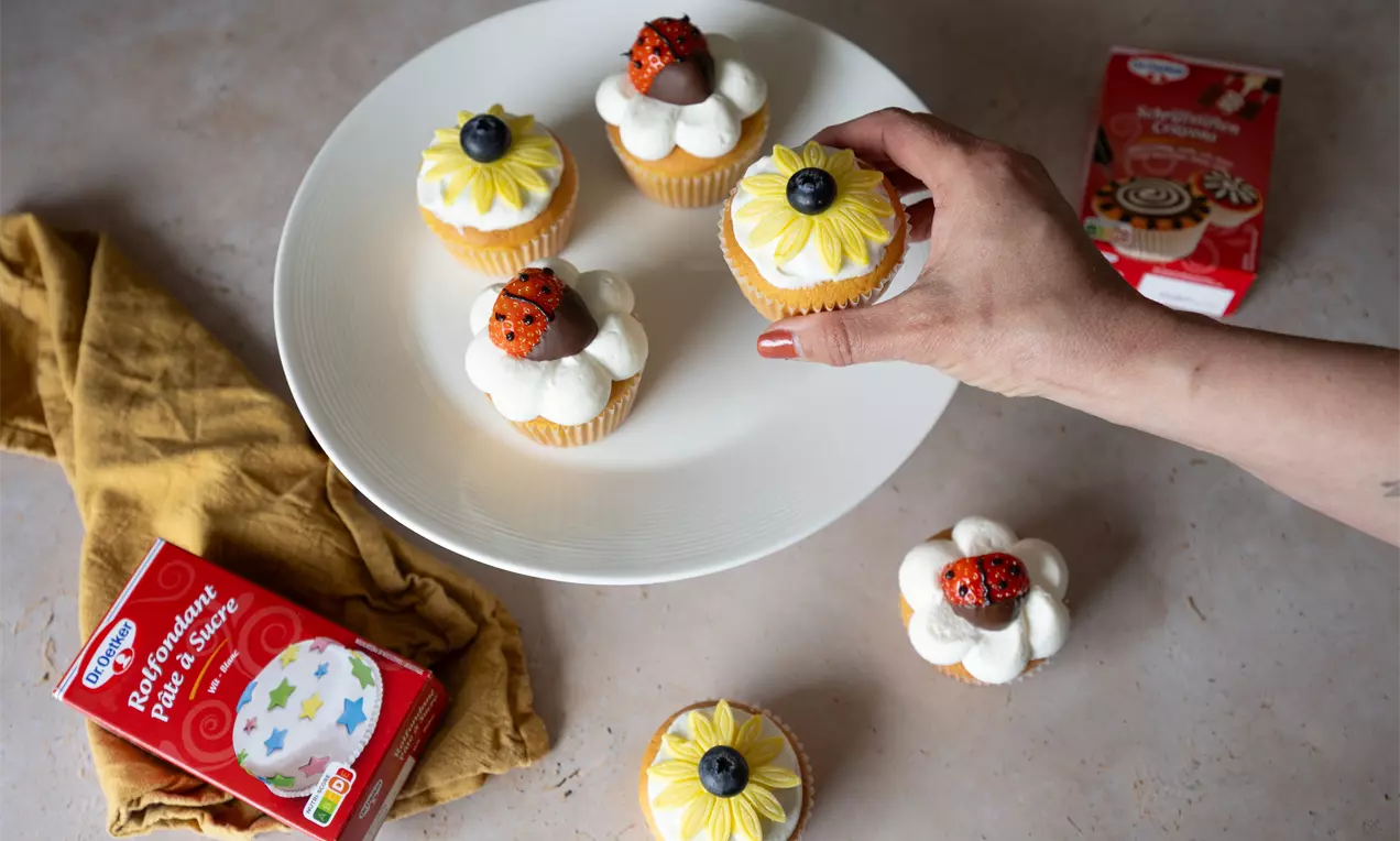 Kleine cupcakes met witte en gele crèmetoefjes, blauwe bessen en chocolaatjes op een wit bord met een hand die een cupcake oppakt