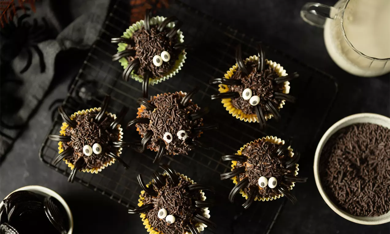 Petits cupcakes enrobés de vermicelles chocolatés, ornés d'yeux en sucre et de pattes en réglisse noire, disposés sur une grille noire