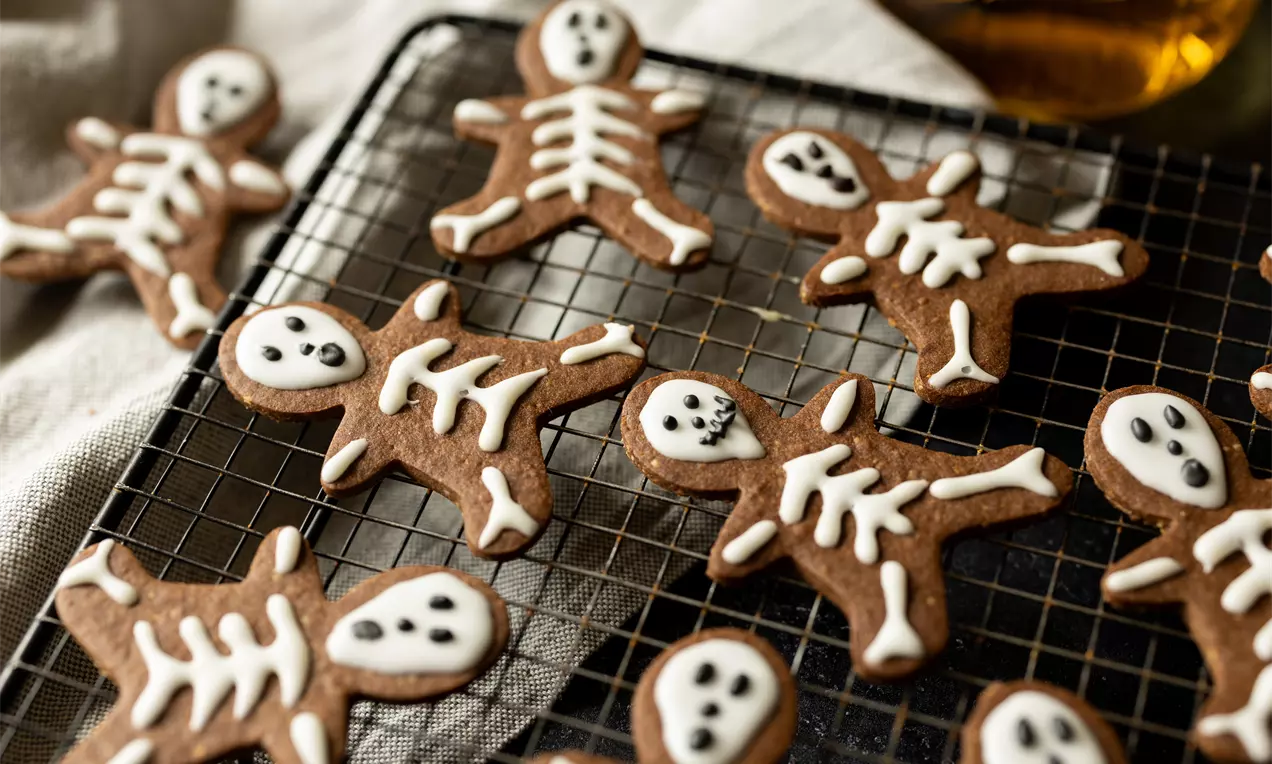 Donkerbruine gingerbread-poppetjes met witte suikerdecoratie in skeletmotief, liggend op een zwarte metalen rooster met een linnen doek ernaast