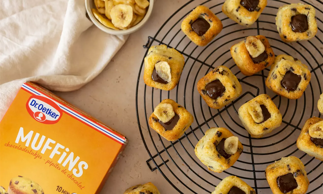 Mini muffins goudgeel en luchtig met stukjes chocola en banaanschijfjes, geplaatst op een metalen koelel, naast een kartonnen verpakking