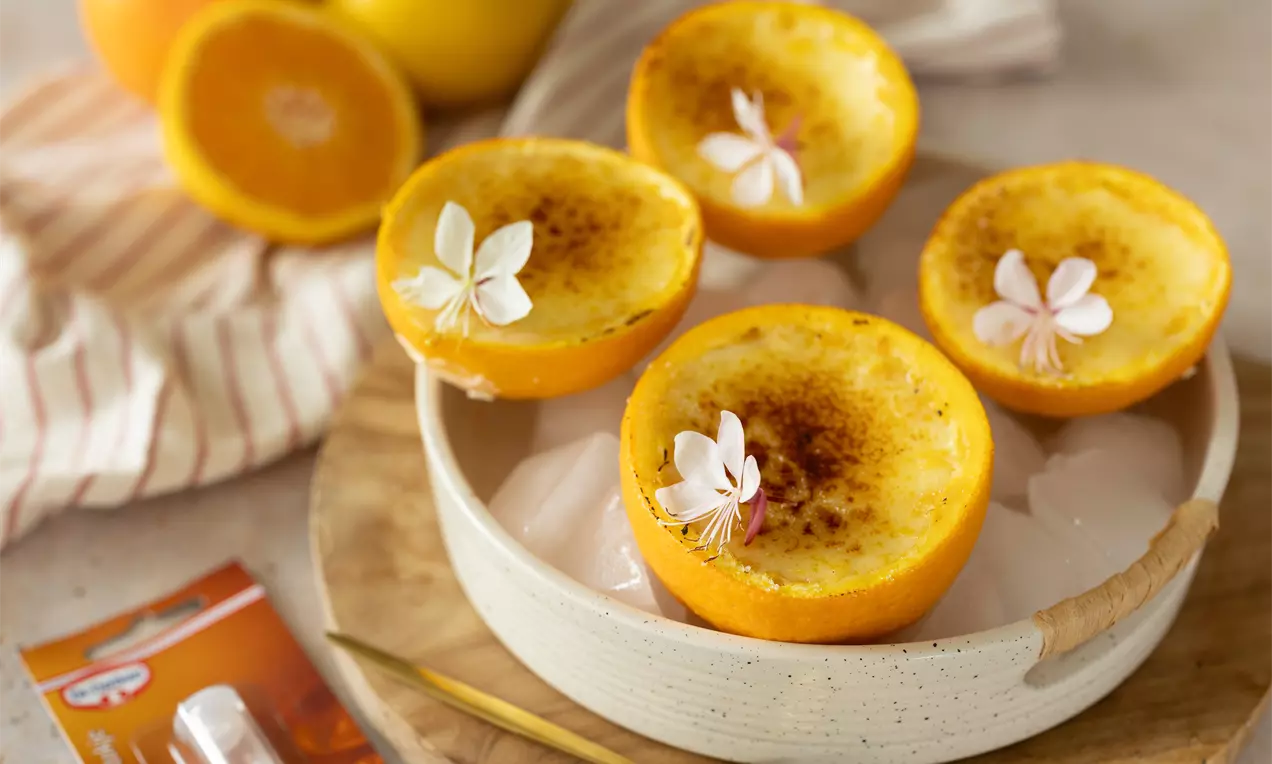 Gehalveerde sinaasappels met gekarameliseerde vulling, versierd met witte bloemen, gepresenteerd in een crèmekleurig schaaltje op houten plank