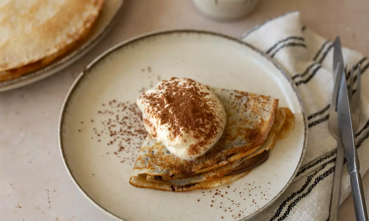 Crêpes dorées pliées en triangles garnies de crème fouettée et saupoudrées de cacao sur une assiette beige avec un napperon à rayures
