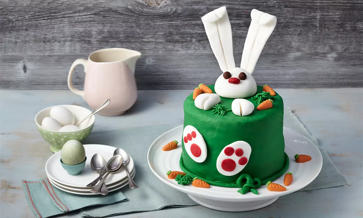 Groene ronde taart met witte fondant oren, witte snoet, rode neus, groene decoraties en kleine wortels op een wit bord