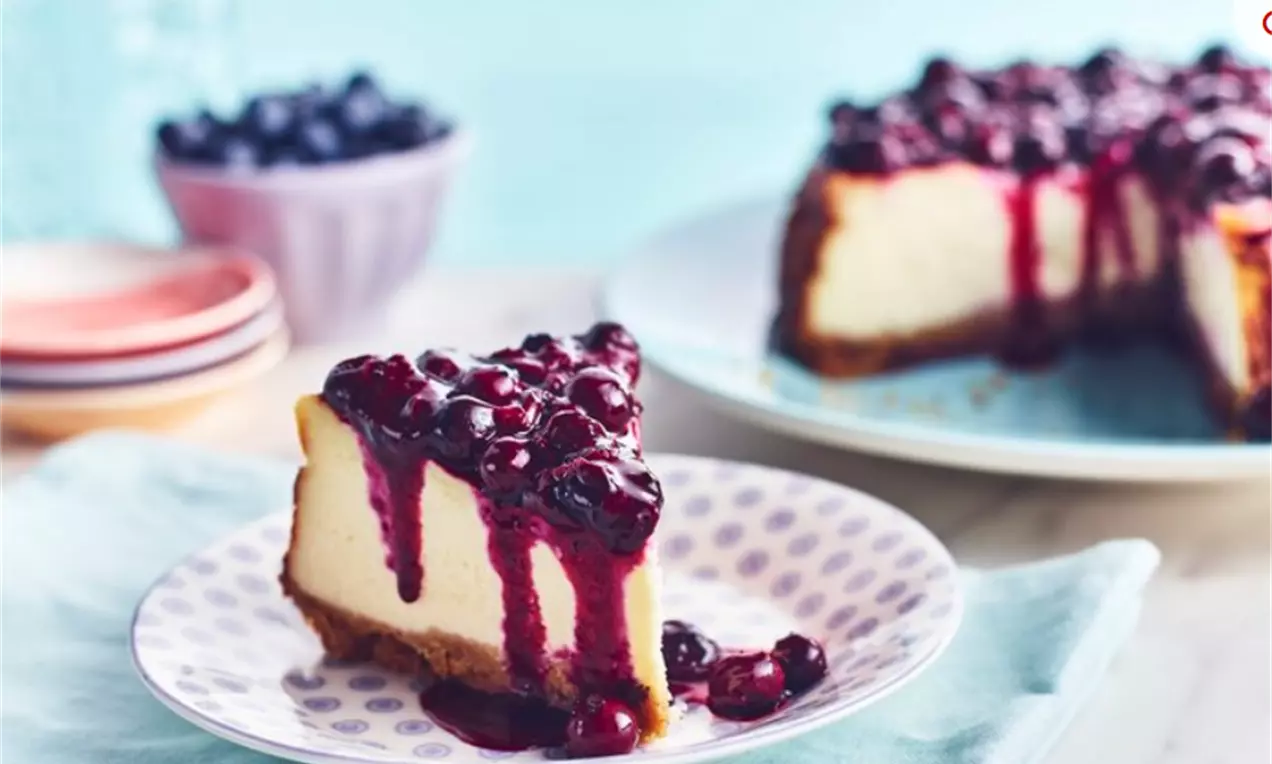 Part de cheesecake à la vanille crémeux sur une base dorée, nappée de compote violette et brillante de myrtilles, posée sur une assiette à motifs