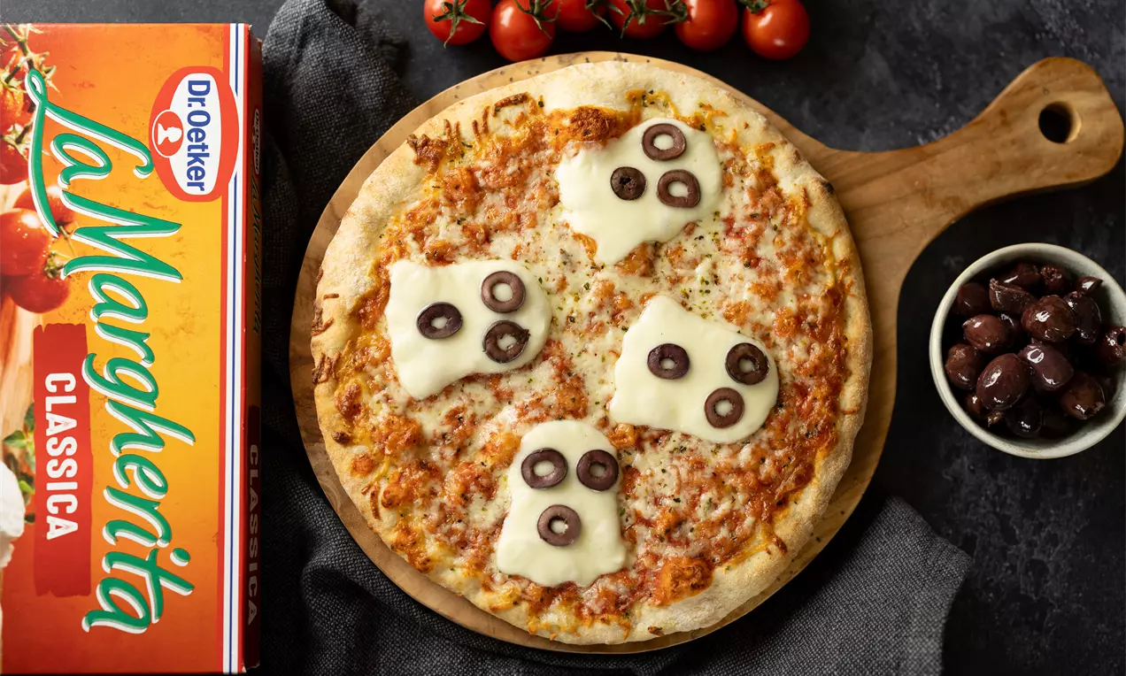 Pizza dorée sur une planche en bois, ornée de tranches de fromage en forme de fantômes et d’olives noires, avec des tomates cerises et un bol d’olives à côté
