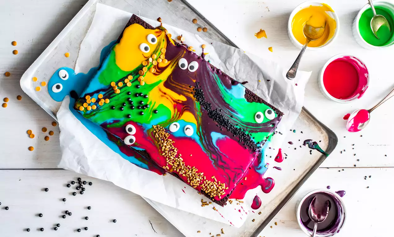 Rechthoekige brownie met felgekleurde glazuurvlekken, suikerogen en gouden en zwarte sprinkels op wit bakpapier op een metalen bakplaat