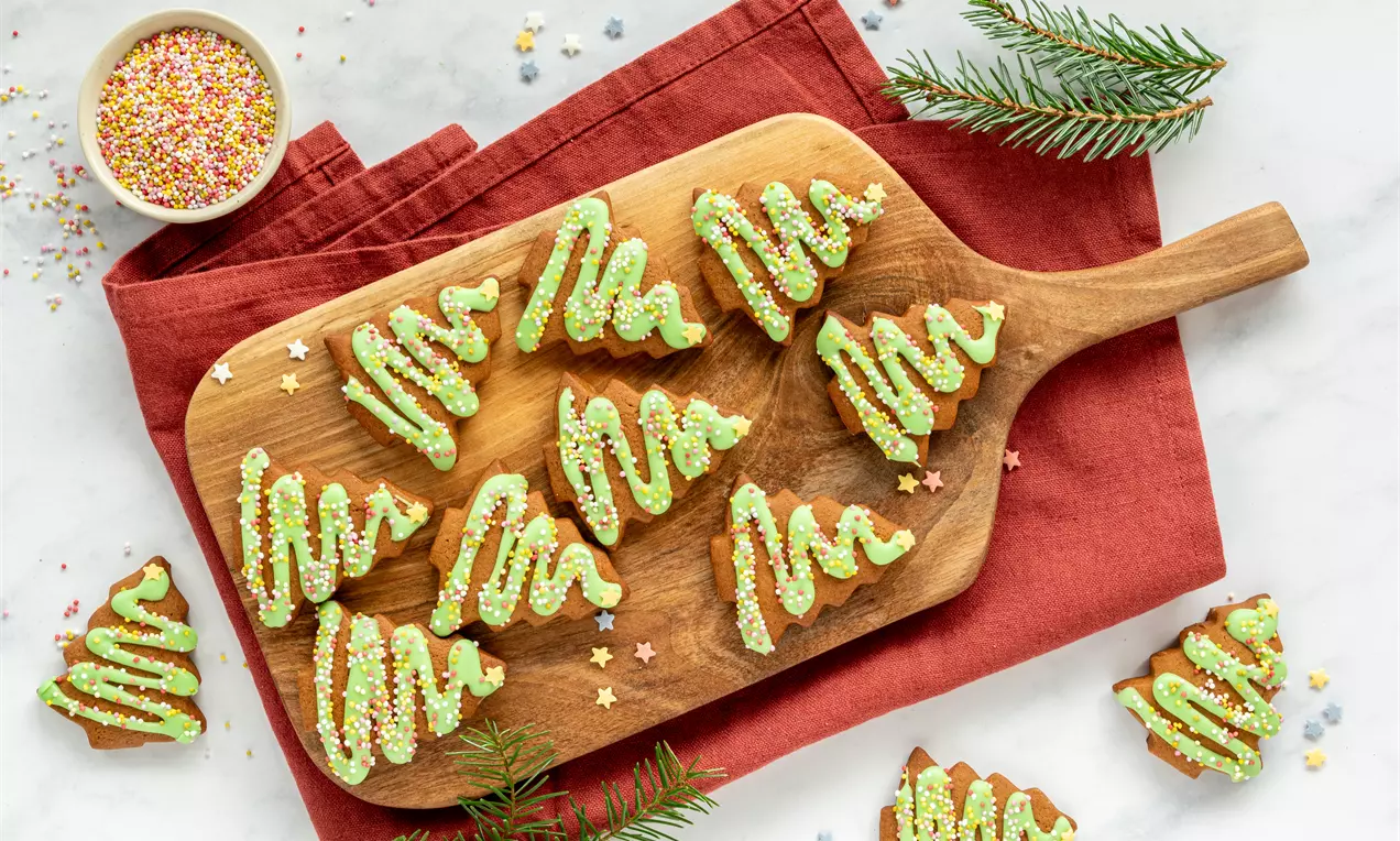 Groene kerstboomkoekjes met glazuur en kleurrijke sprinkles, netjes gerangschikt op een houten plank met rode stof en dennentakken