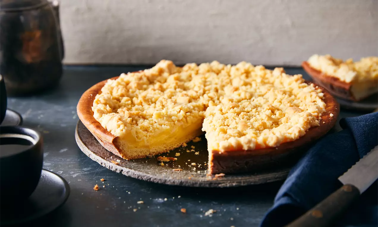 Tarte ronde dorée avec une base moelleuse, garnie de crème pâtissière jaune et d’un crumble croustillant, sur une assiette en pierre