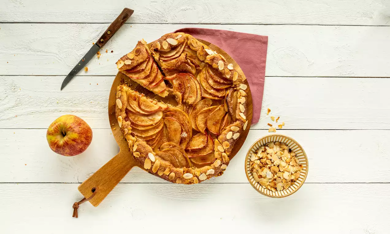 Galette met dunne plakken goudbruine appel, omrand met een krokant deeg en geschaafde amandelen op een houten plank met roze doek