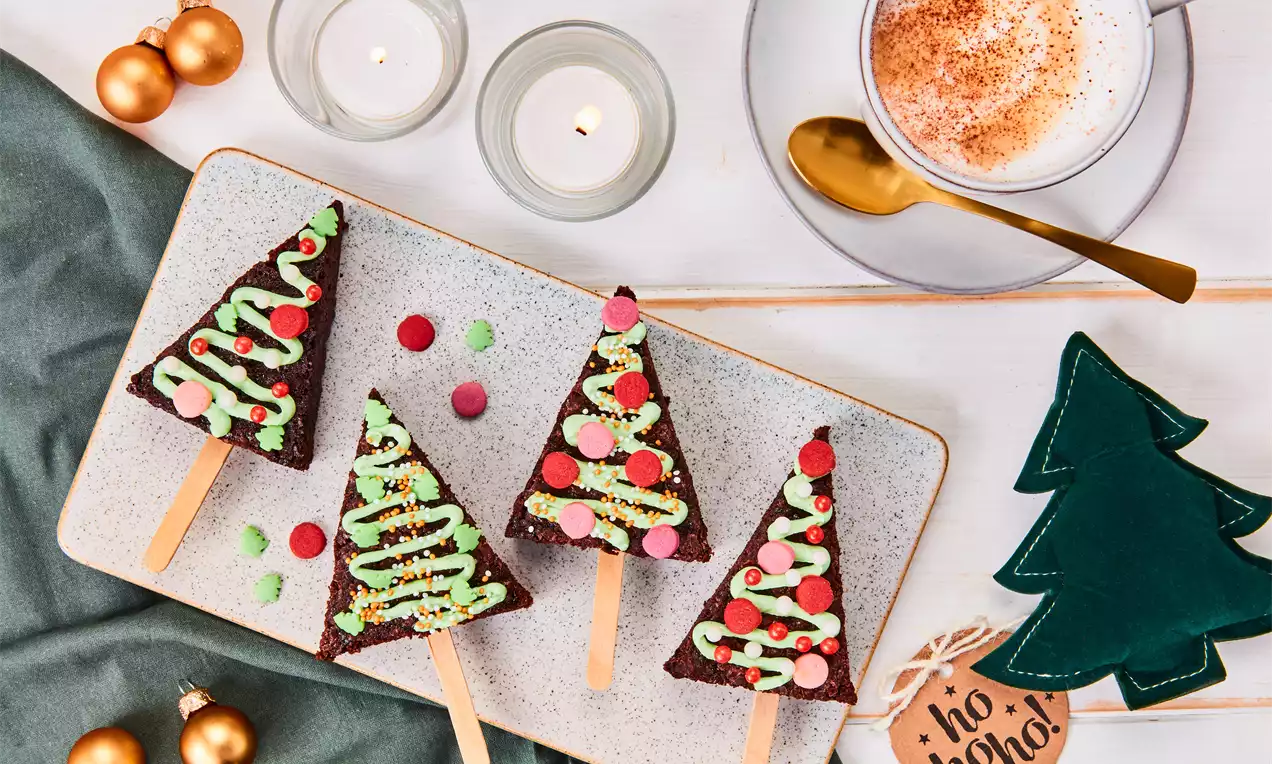 Driehoekige kerstbrownies op houten stokjes, versierd met groene glazuur en gekleurde sprinkles, liggend op een wit bord met kerstdecoratie