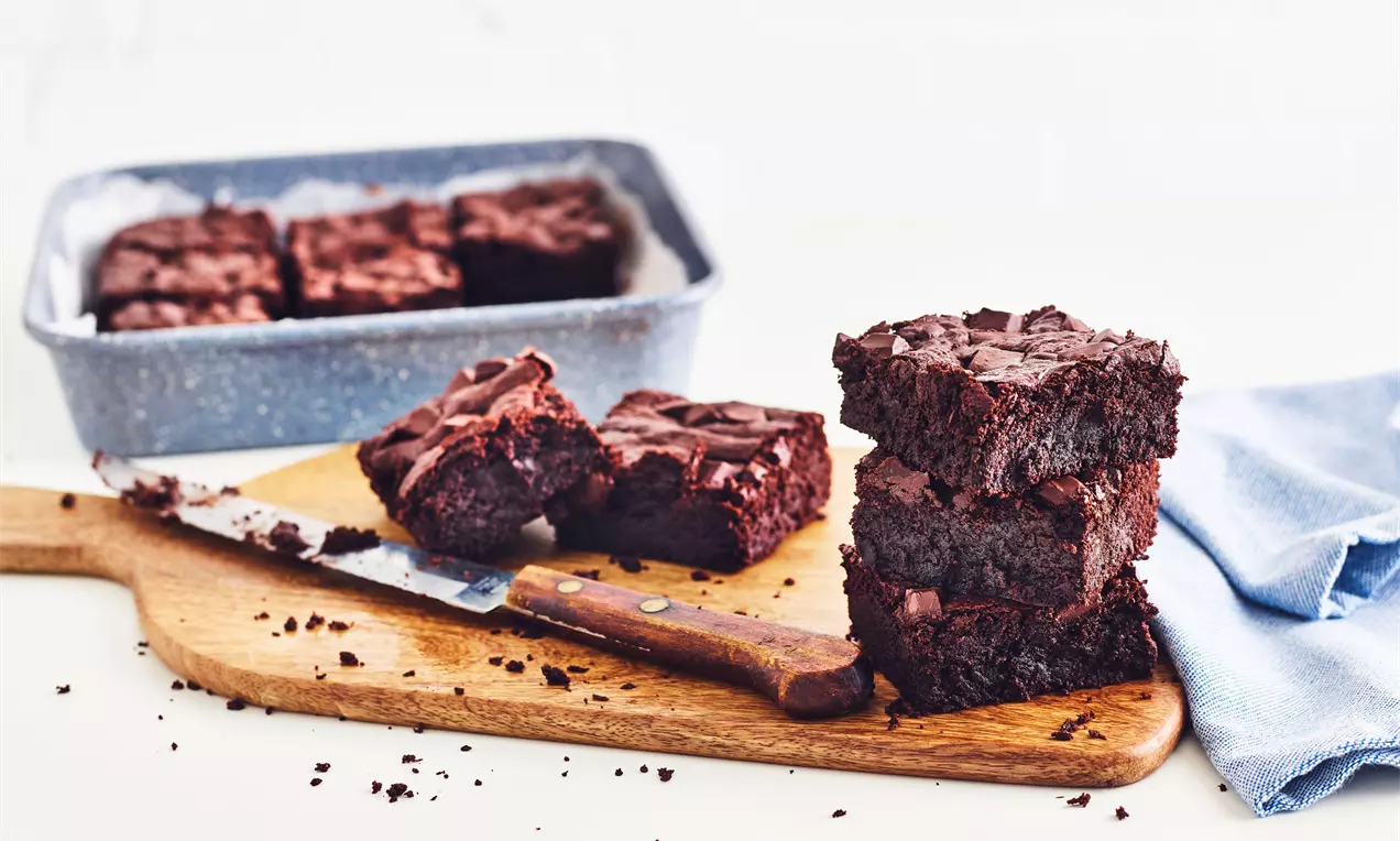 Donkerbruine vegan brownies met een zachte, kruimelige textuur op een houten plank naast een mes, een blauwe doek en een bakvorm