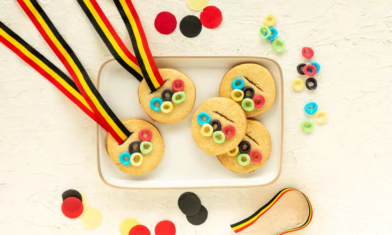 Goudbruine ronde koekjes met kleurrijke ringdecoraties en linten in zwart, geel en rood, gerangschikt op een wit bord