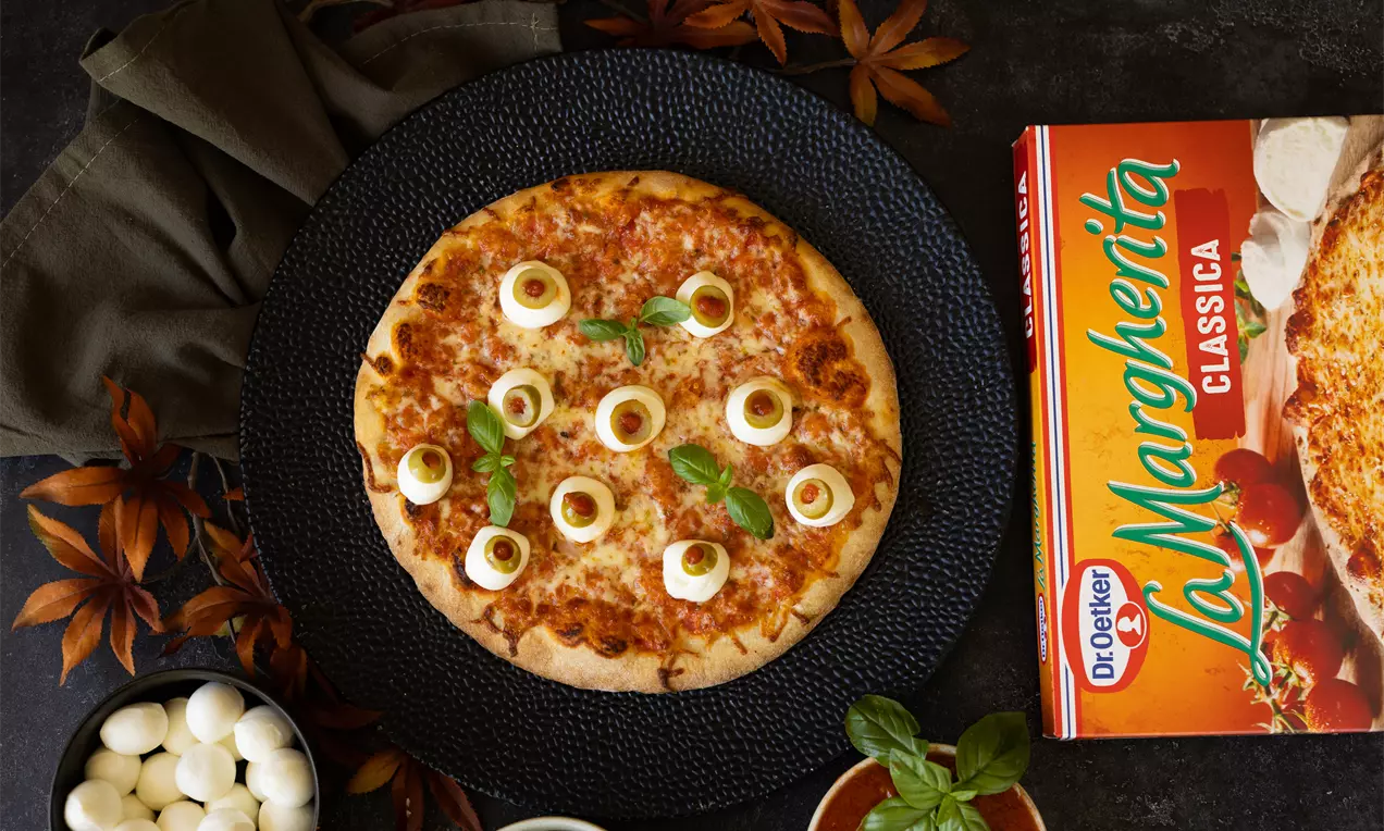 Pizza ronde dorée recouverte de mozzarella fondante, rondelles d’olives et feuilles de basilic, servie sur une assiette noire.