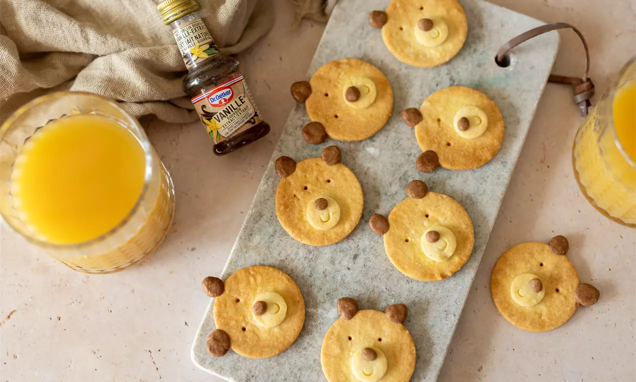 Biscuits dorés en forme de visages d'ours avec des oreilles en chocolat et un nez vanillé sur une planche de marbre, entourés de jus d'orange