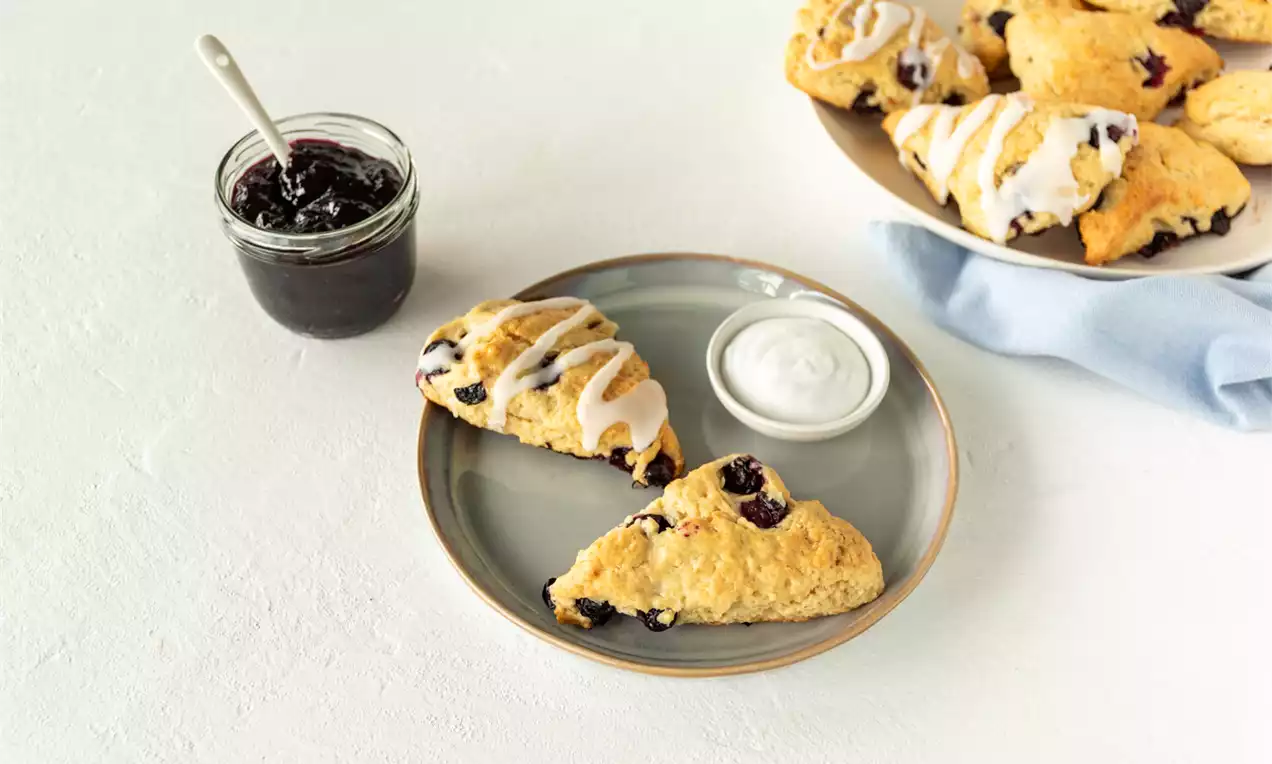 Lichtbruine driehoekige vegan scones met glazuur en blauwe bessen op een grijze schaal met jam en room, naast een witte schaal met scones
