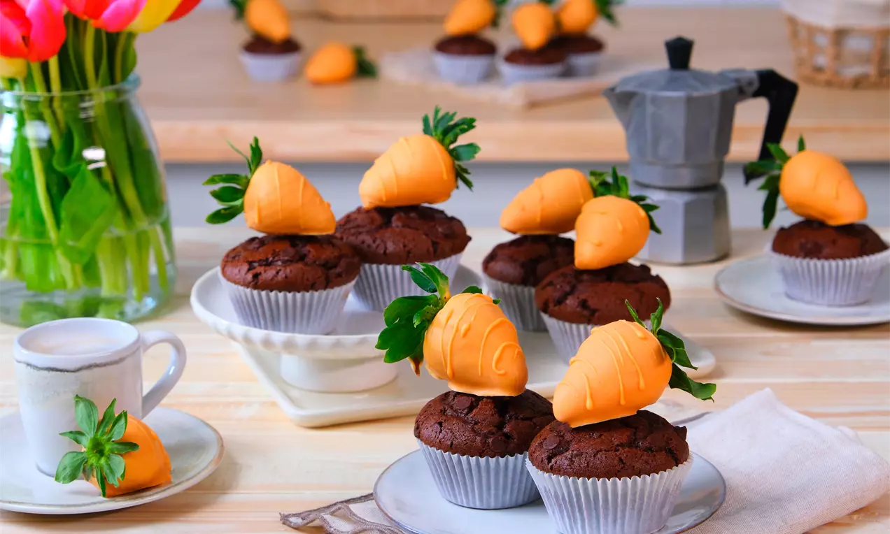 Chocolade muffins met witte papieren vormpjes, gedecoreerd met glanzende oranje aardbeien in de vorm van worteltjes met groene blaadjes