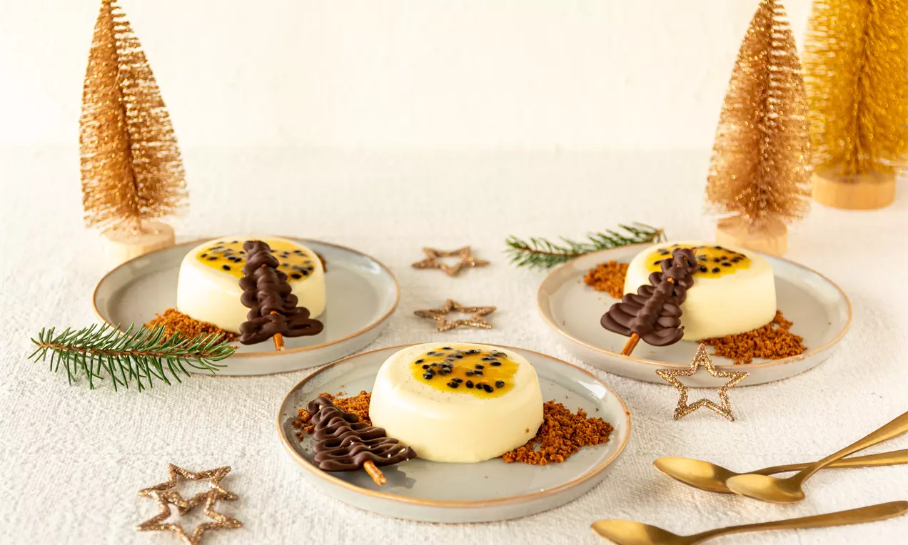 Romige witte panna cotta gegarneerd met gele passievrucht op een bord met chocoladekerstboom en koekkruimels tussen gouden kerstdecoraties