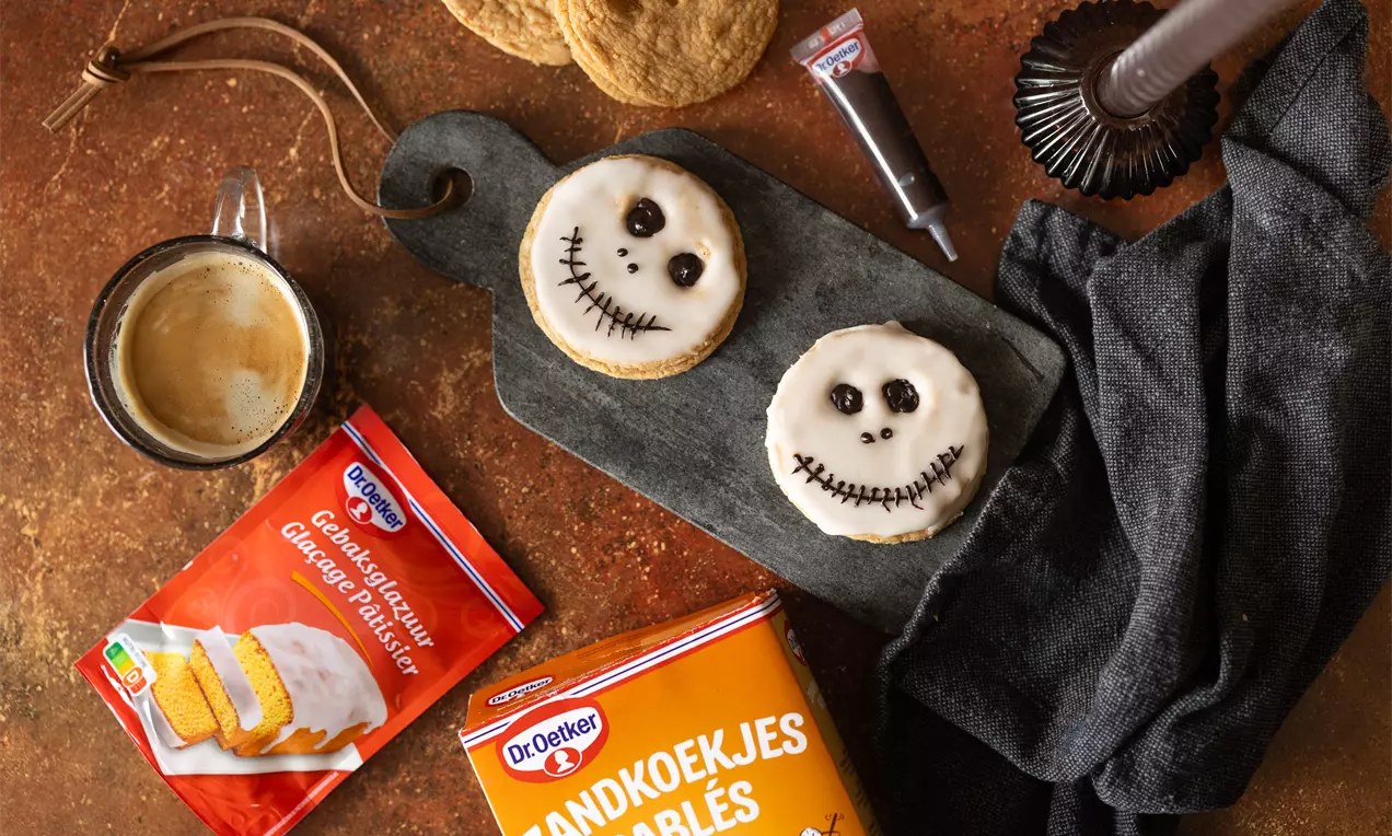 Cookies ronds recouverts de glaçage blanc avec des visages dessinés en chocolat sur une planche noire accompagnés d'un café et d'emballages