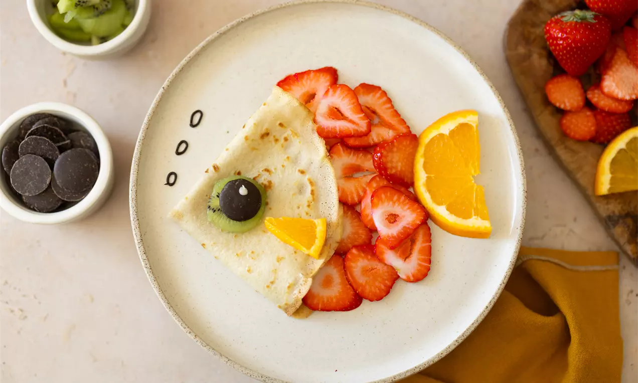 Een dunne pannenkoek met fruitdecoratie in de vorm van een vogel, gemaakt van aardbeien, sinaasappel, kiwi en chocolade op een wit bord