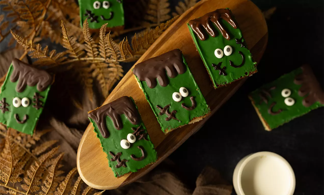 Groene rechthoekige traktaties met gezichtjes, bedekt met donkere chocolade bovenop, gepresenteerd op houten plank tussen herfstbladeren