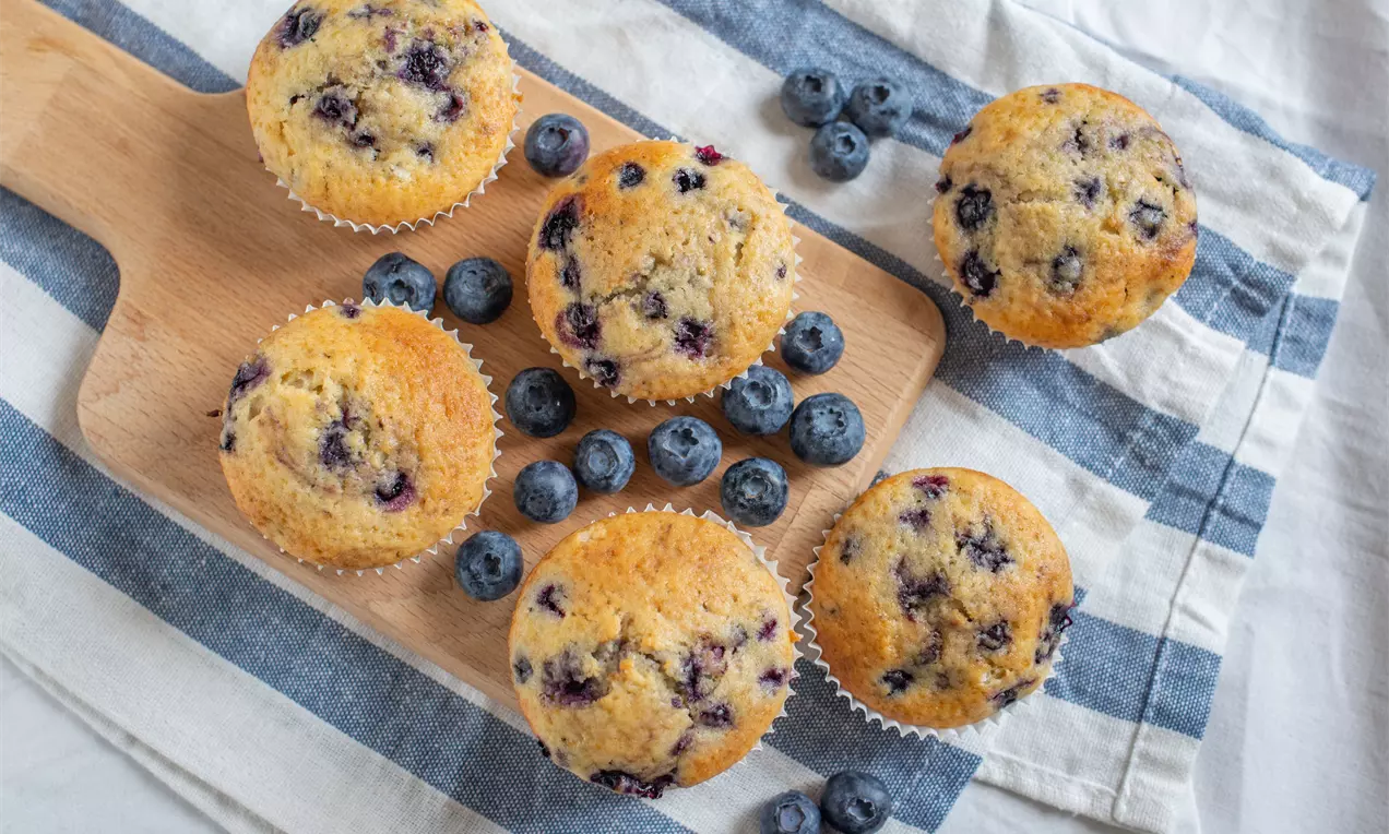 Muffins dorés parsemés de myrtilles fraîches sur une planche en bois claire, entourés de myrtilles sur un linge rayé blanc et bleu