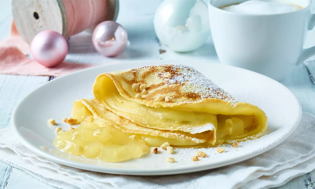 Goudgele gevulde pannenkoek met vulling die uitloopt, bestrooid met poedersuiker en nootjes op een wit bord, met winterdecoratie op de achtergrond