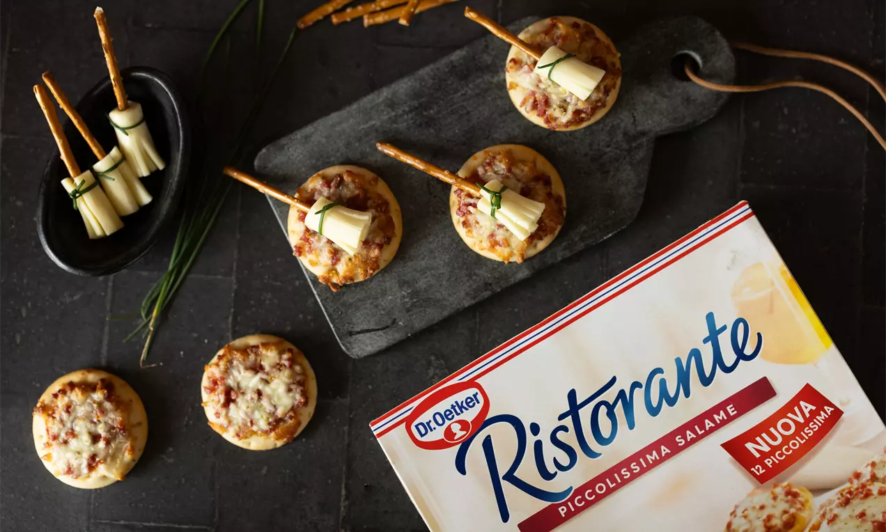 Mini pizzas dorées garnies de fromage fondu et éclats de salami rouge, disposées sur une planche sombre avec bâtonnets de bretzels et oignons nouveaux