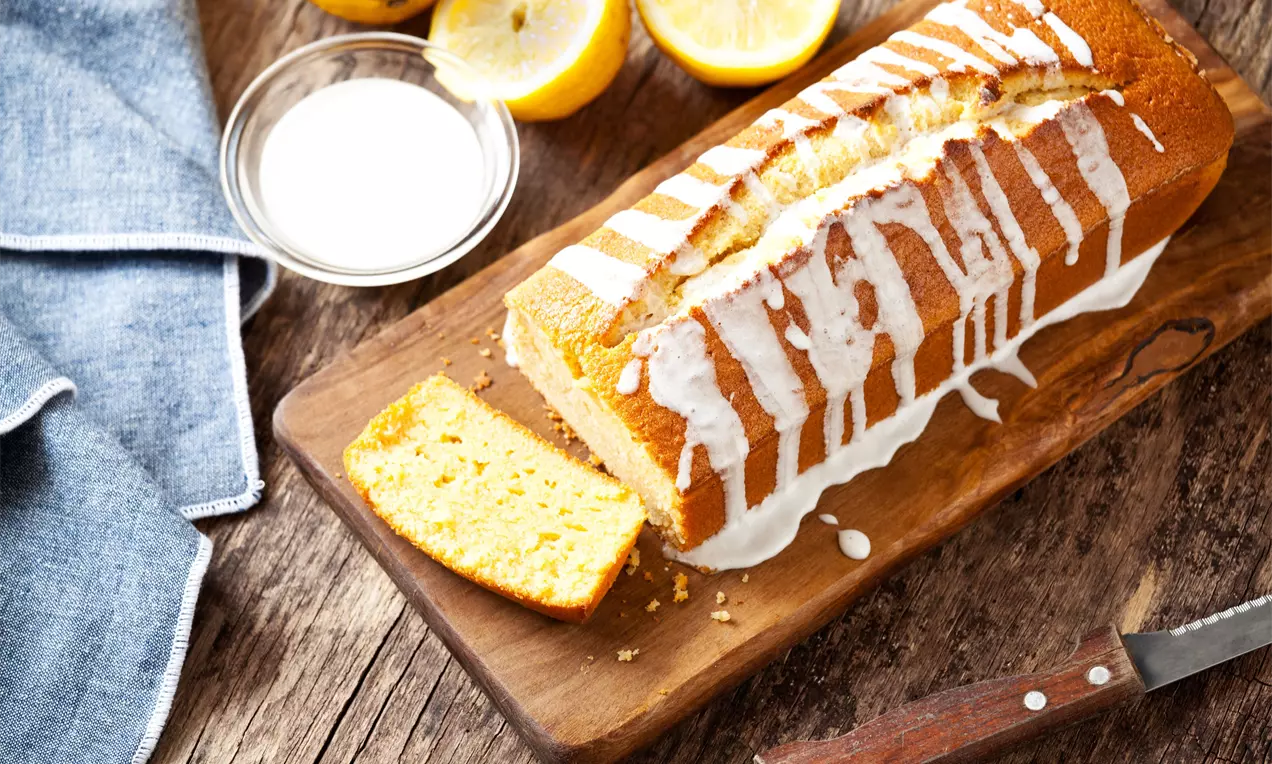 Cake doré au citron à la texture moelleuse, nappé de glaçage blanc coulant, présenté sur une planche en bois avec un couteau et des citrons jaunes coupés