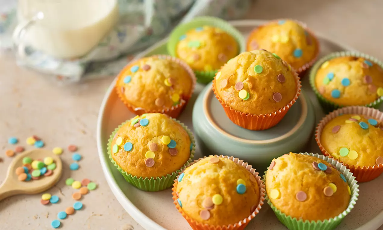Muffins dorés moelleux dans des caissettes colorées, décorés de confettis sucrés multicolores, disposés sur une assiette beige.