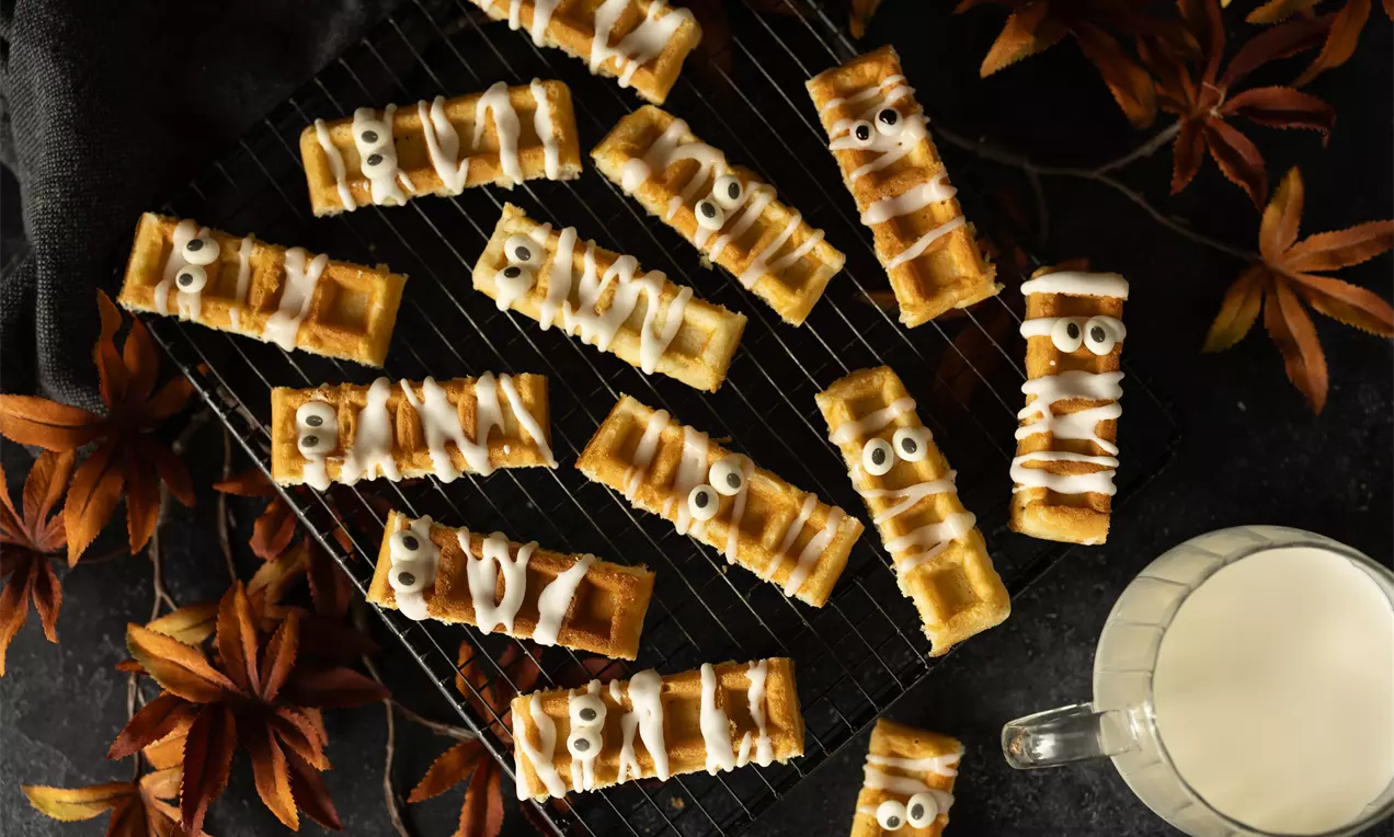 Rechthoekige koekreepjes met witte glazuurstrepen en suikerogen, uitgestald op een zwarte roosterbakplaat met herfstbladeren eromheen