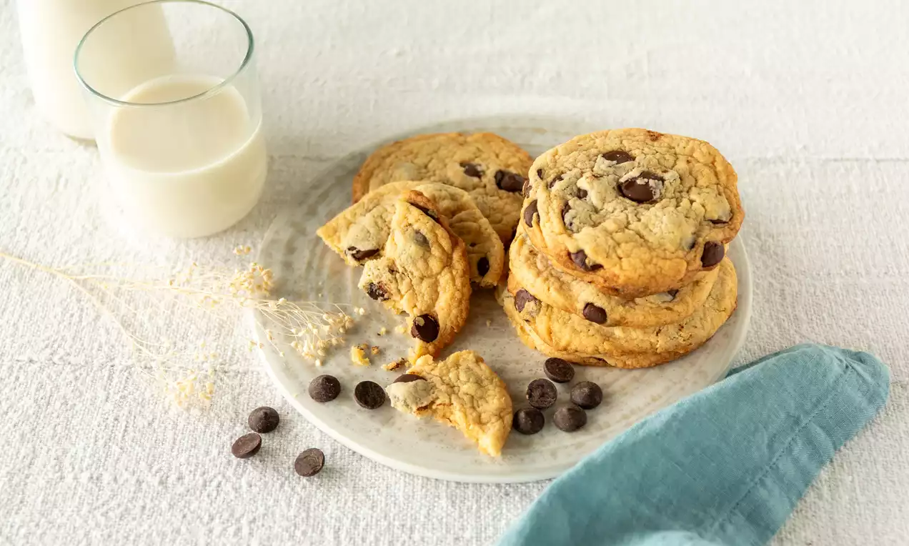 Ronde goudbruine vegan chocolate chip cookies met knapperige randen op een wit bord, omringd door chocoladestukjes en glazen melk