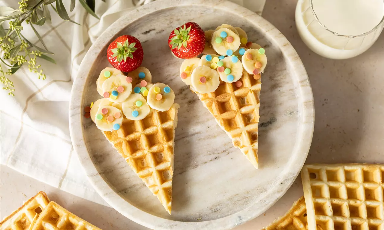 Wafelpunten gegarneerd met plakjes banaan, kleurrijke strooisels en aardbeien, gepresenteerd op een wit marmeren bord met decoratieve takjes