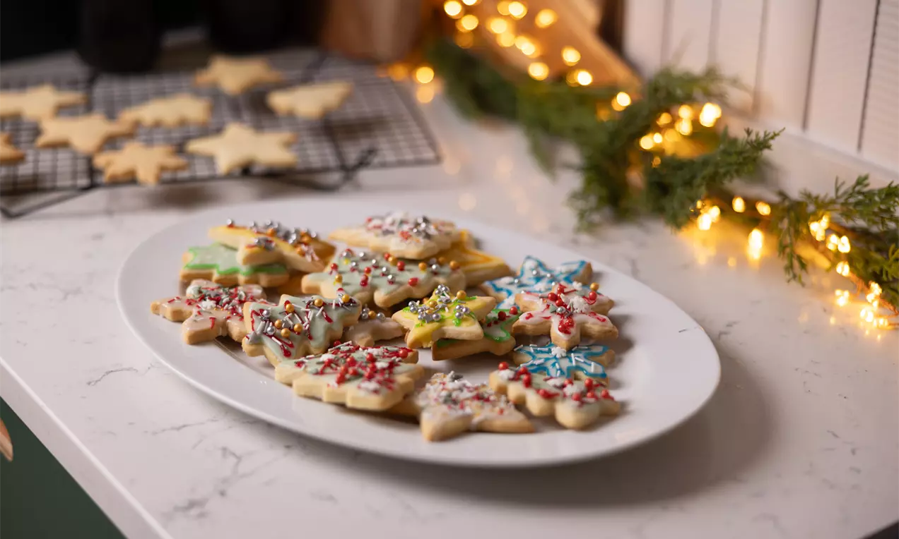 Op een wit bord liggen kerstkoekjes in stervorm, rijkelijk gedecoreerd met kleurrijke glazuur en sprinkles, omringd door lichte kerstdecoratie